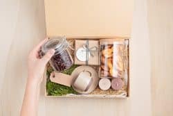 A woman's hand placing a mason jar full of coffee beans into a box with an assortment of candles, soaps, and other gift items.